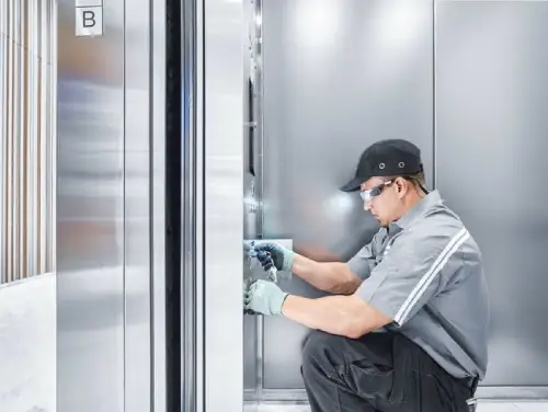 Ліфтер виконує технічне обслуговування при відчинених дверях ліфта, в захисних окулярах і кепці.